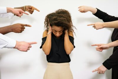 Une jeune femme aux cheveux bouclés semble affligée alors que plusieurs mains pointent des doigts accusateurs vers elle.